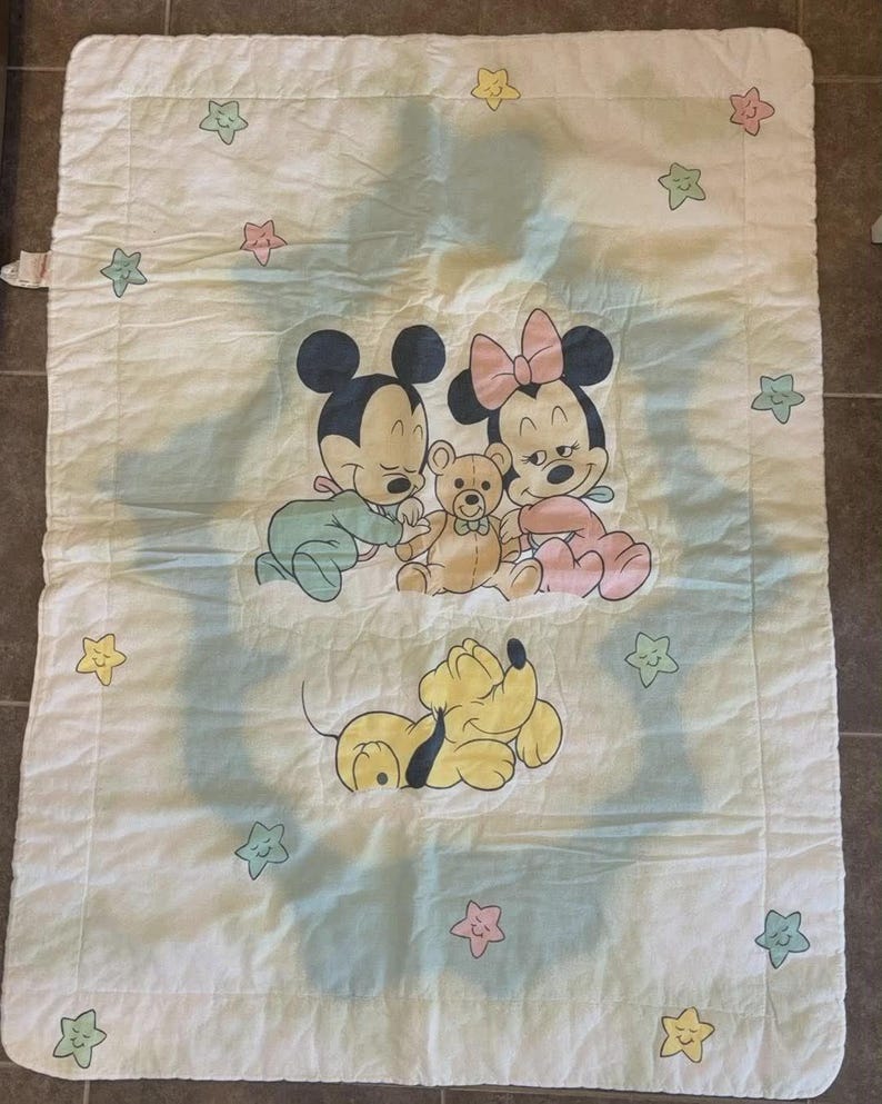 Puede incluir: Una manta rectangular blanca para beb&eacute; con una ilustraci&oacute;n central de los beb&eacute;s Mickey y Minnie Mouse abrazando un oso de peluche, con Pluto durmiendo debajo. El borde est&aacute; decorado con peque&ntilde;as estrellas de colores pastel.