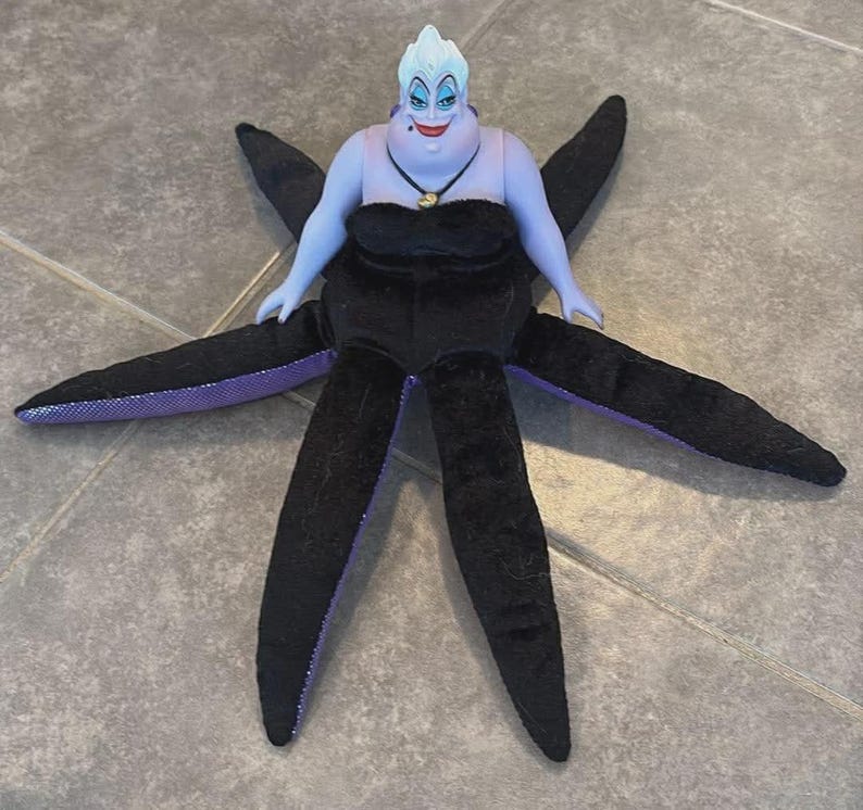 May include: A plush doll of Ursula, the sea witch, from Disney's The Little Mermaid. The doll features a light blue face and arms, a black dress, and six black tentacles with purple undersides. A gold necklace adorns her neck.