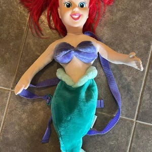 May include: A plush Ariel backpack with a purple seashell bra, a green tail, and a light blue tail fin. The backpack has purple straps.
