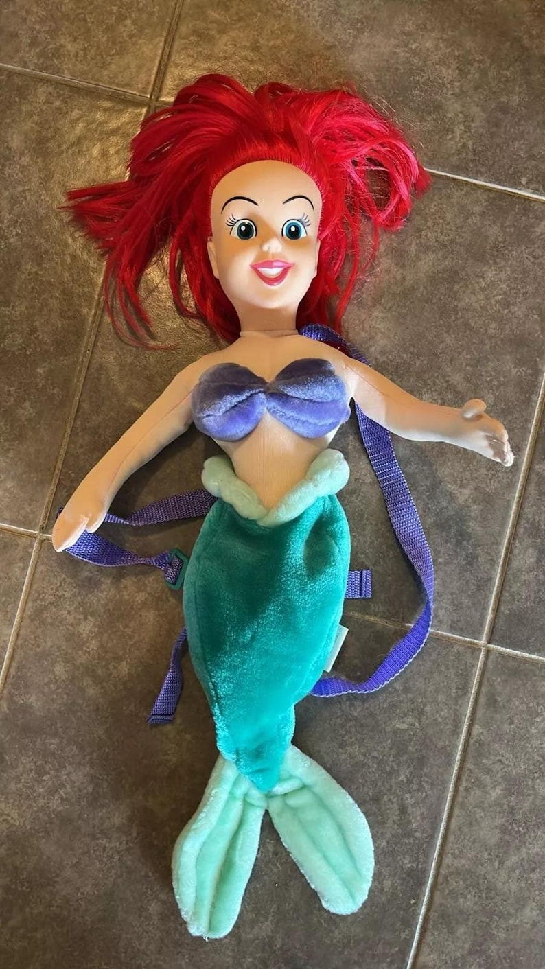 May include: A plush Ariel backpack with a purple seashell bra, a green tail, and a light blue tail fin. The backpack has purple straps.