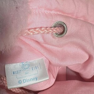 May include: A close-up of a pink drawstring bag with a fluffy, light pink trim. The bag has a silver grommet and a pink drawstring. A white tag with the Disney logo is visible.
