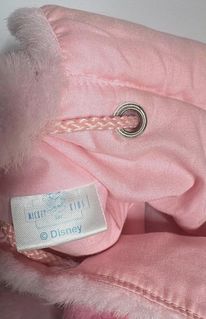 May include: A close-up of a pink drawstring bag with a fluffy, light pink trim. The bag has a silver grommet and a pink drawstring. A white tag with the Disney logo is visible.