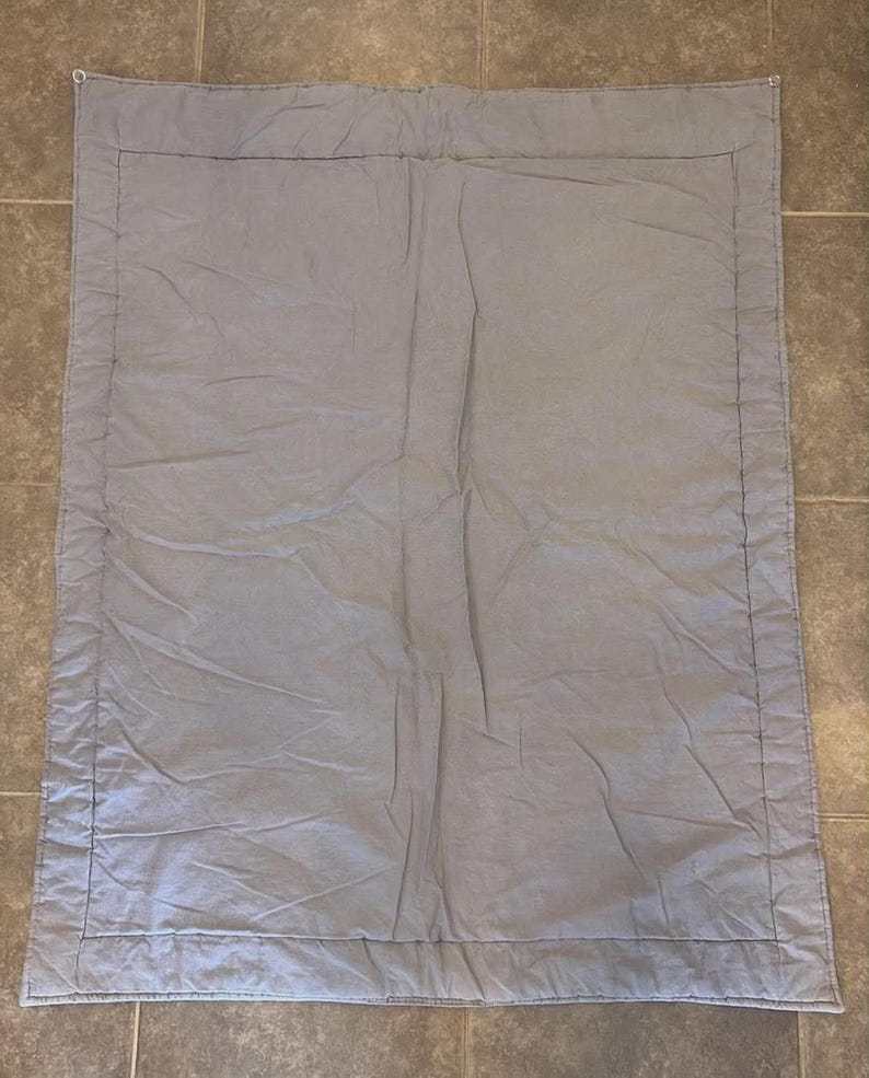 May include: A rectangular light gray quilt featuring a solid color design with a stitched border frame. The fabric has a soft, slightly wrinkled texture throughout the surface, with visible stitching lines defining the central panel and edges.