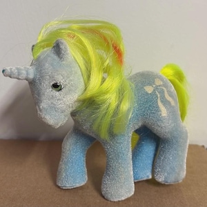 Könnte beinhalten: Ein hellblaues Plüsch-Einhorn-Spielzeug mit gelber Mähne und Schweif sowie einem weißen Horn. Das Einhorn hat ein weißes Schleifendesign an der Seite und grüne Augen. Die Mähne hat orangefarbene Streifen.