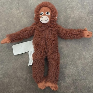 May include: A soft toy orangutan with brown fur and a lighter brown face, arms outstretched. The toy features embroidered facial details, including large eyes and a smiling mouth. A white tag is attached.