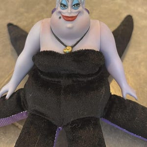 May include: A plush doll of Ursula, the sea witch from Disney's The Little Mermaid. The doll has a purple face and arms, white hair, and a black dress. She wears a shell necklace and has black tentacles.