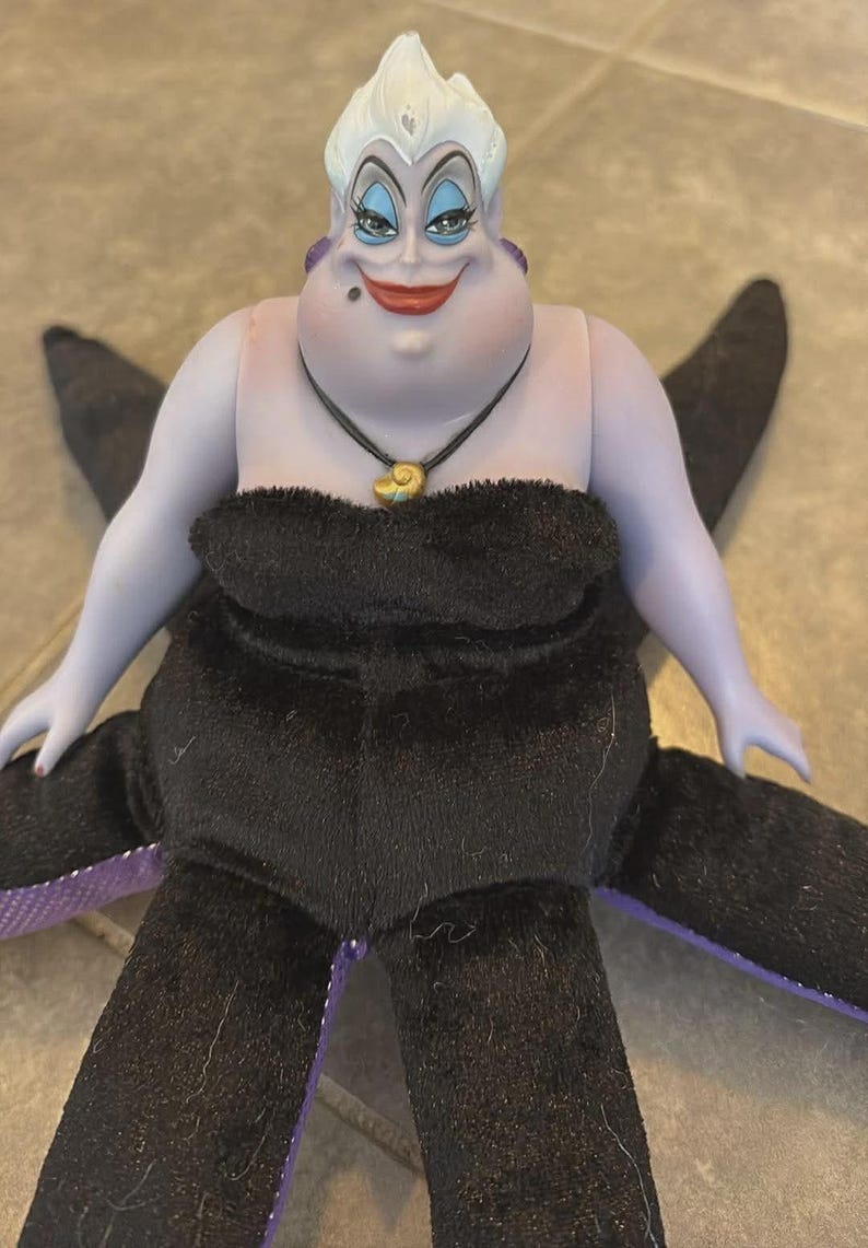 May include: A plush doll of Ursula, the sea witch from Disney's The Little Mermaid. The doll has a purple face and arms, white hair, and a black dress. She wears a shell necklace and has black tentacles.