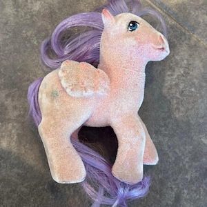 Puede incluir: Un juguete de peluche rosa vintage con pelo y cola morados. El pony tiene ojos azules, una pequeña ala rosa a un lado y un cuerpo rosa claro. El juguete está de pie sobre sus cuatro patas.