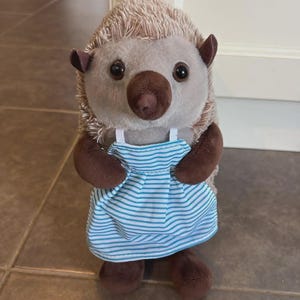 Pode incluir: Um brinquedo de pelúcia de ouriço com corpo castanho claro, braços e pés castanhos escuros e um vestido listrado azul claro e branco. O ouriço tem olhos grandes e escuros e um nariz castanho.