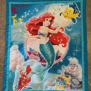 May include: A rectangular quilt featuring a vibrant underwater scene with a red-haired mermaid, a yellow fish, a red crab, a white-haired king, and a purple-skinned sea witch. The design includes blue water, bubbles, and a pearl border.