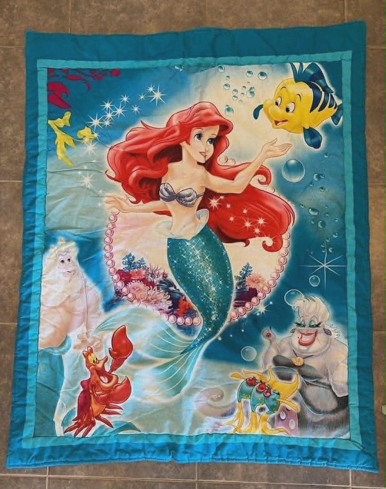 May include: A rectangular quilt featuring a vibrant underwater scene with a red-haired mermaid, a yellow fish, a red crab, a white-haired king, and a purple-skinned sea witch. The design includes blue water, bubbles, and a pearl border.
