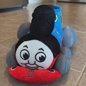 May include: A plush toy shaped like a blue steam locomotive with a black smokestack, a white face featuring large black eyes and a smiling mouth, and a red front bumper. The toy has four grey wheels and blue accents on the sides of the body.
