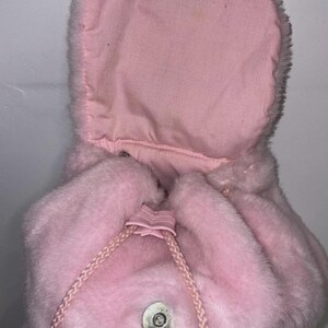 May include: A pink, plush backpack with a drawstring closure and a flap that secures with a snap. The backpack has a soft, furry texture and a matching pink drawstring. The hood is open, revealing a lighter pink interior.