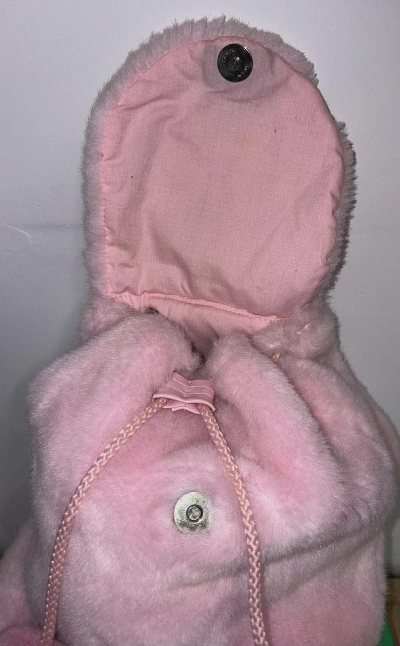 May include: A pink, plush backpack with a drawstring closure and a flap that secures with a snap. The backpack has a soft, furry texture and a matching pink drawstring. The hood is open, revealing a lighter pink interior.