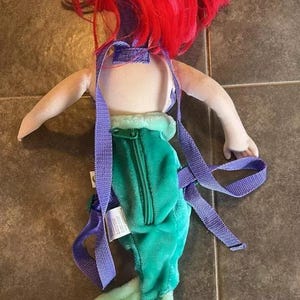 May include: A plush Ariel backpack with purple straps. The backpack is shaped like Ariel from The Little Mermaid, with a green tail and red hair.