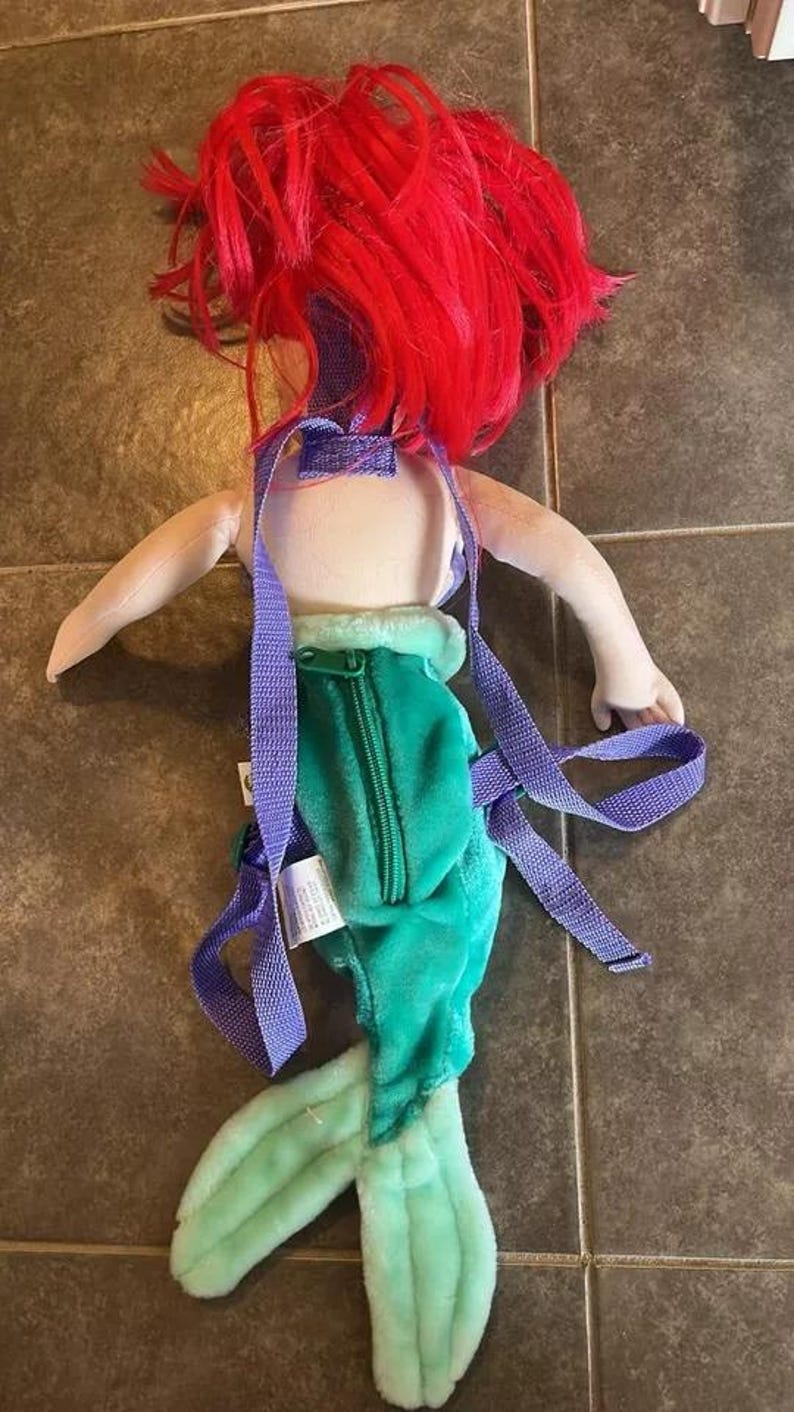 May include: A plush Ariel backpack with purple straps. The backpack is shaped like Ariel from The Little Mermaid, with a green tail and red hair.