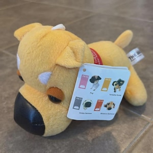 May include: A yellow plush dog toy with a black nose and brown accents. The toy has a red collar and a tag with images of different dog breeds, including a Pug, Yorkshire Terrier, Golden Retriever, and Miniature Dachshund.