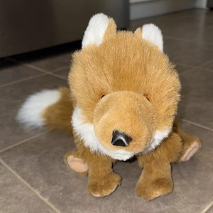 Puede incluir: Un juguete de peluche de zorro con pelaje marrón, detalles blancos en las orejas y el pecho, y ojos negros. El zorro está sentado en un suelo de baldosas.