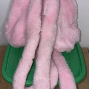 May include: A fluffy, light pink faux fur hat with long, dangling ear-like extensions. The hat is made of soft, plush material, and is designed to resemble bunny ears. The hat is resting on a green plastic container.