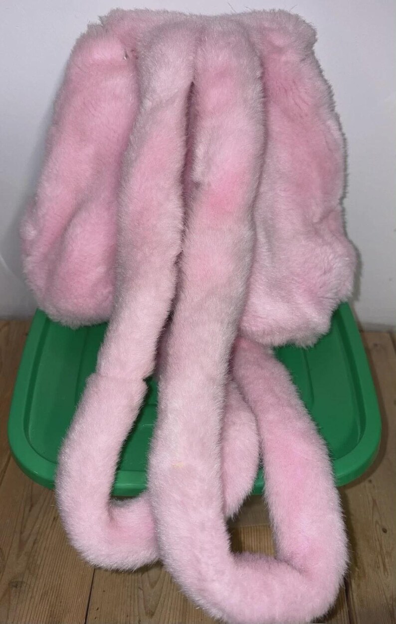 May include: A fluffy, light pink faux fur hat with long, dangling ear-like extensions. The hat is made of soft, plush material, and is designed to resemble bunny ears. The hat is resting on a green plastic container.