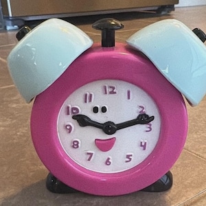 May include: A pink twin-bell alarm clock with a white face featuring purple numbers, black clock hands, and a smiling face design. The clock has two light blue bells on top, a black top button, and black feet, creating a colorful, functional timepiece.