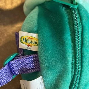 May include: A green plush backpack with a zipper closure and a purple strap. The backpack has a Disney's The Little Mermaid label attached to it. The label is white with blue and yellow text. The text on the label reads "Disney's The Little Mermaid" and "Disney".