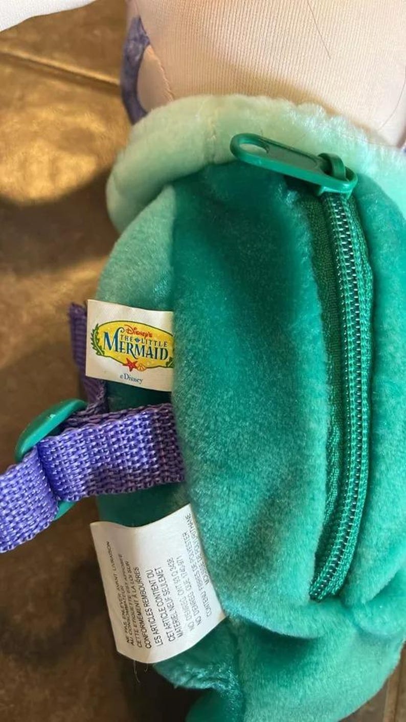 May include: A green plush backpack with a zipper closure and a purple strap. The backpack has a Disney's The Little Mermaid label attached to it. The label is white with blue and yellow text. The text on the label reads "Disney's The Little Mermaid" and "Disney".