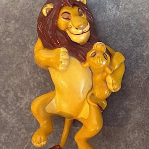 Puede incluir: Figura amarilla y marrón de un león sosteniendo un cachorro de león más pequeño. El león tiene una melena marrón y una expresión sonriente. El cachorro también es amarillo y tiene una expresión similar. La figura es probablemente un objeto de colección o decorativo.