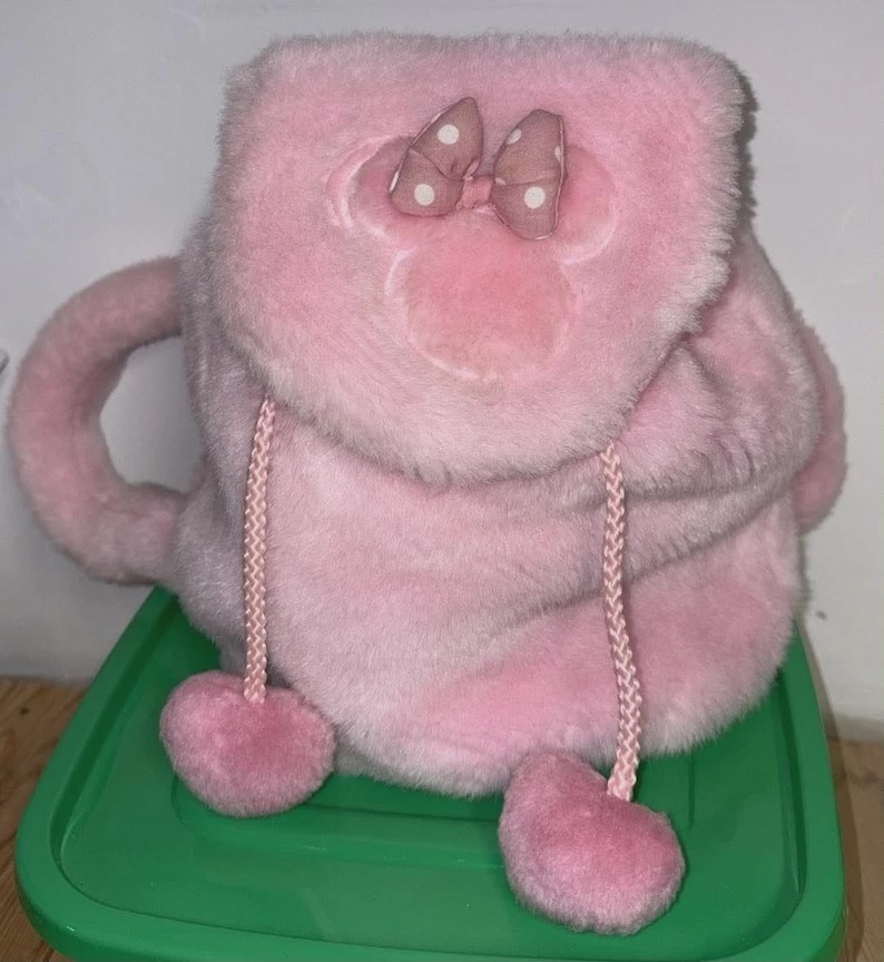May include: A plush pink backpack with a Minnie Mouse design. The backpack features a bow with white polka dots, a drawstring closure, and heart-shaped accents. The bag has a handle and is made of soft, fuzzy material.