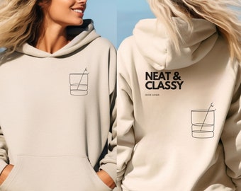 Pode incluir: Dois casacos de moletom bege com um gráfico de um copo com um canudo neles. Um casaco tem o texto "Neat & Classy" com as palavras "Iron Arms" abaixo.