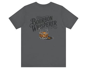 Könnte beinhalten: Ein dunkelgraues T-Shirt mit dem Text "Bourbon Whisperer" in einer stilisierten Schriftart. Unter dem Text ist ein Glas mit bernsteinfarbener Flüssigkeit abgebildet.