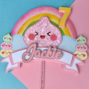 May include: A cake topper featuring a rainbow, a yellow number 7, and a central pink character with a smiling face. A white banner with the name Jackie in pink glitter script sits below. Stacks of small colorful faces flank the sides on a clear stick.