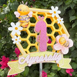 Puede incluir: Adorno para tarta amarillo y rosa con diseño de panal, un número 1 rosa brillante y el nombre "Aynaraliz". El adorno presenta abejas de dibujos animados, margaritas y una pancarta. Ideal para una celebración de primer cumpleaños.
