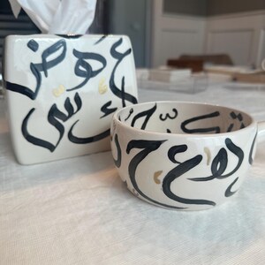 May include: A white ceramic tissue box and matching mug with black calligraphy and gold accents. The tissue box has a square shape with a top opening for tissues. The mug has a handle and the same calligraphy design.