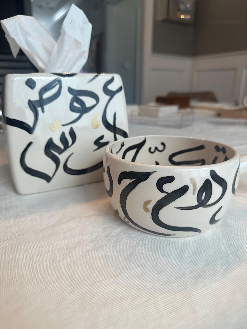 May include: A white ceramic tissue box and matching mug with black calligraphy and gold accents. The tissue box has a square shape with a top opening for tissues. The mug has a handle and the same calligraphy design.