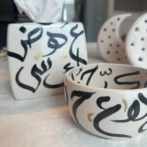 May include: A white ceramic tissue box and bowl set with black and gold calligraphy. The tissue box is rectangular with a square opening for tissues. The bowl is round. Two decorative moon-shaped objects are in the background.