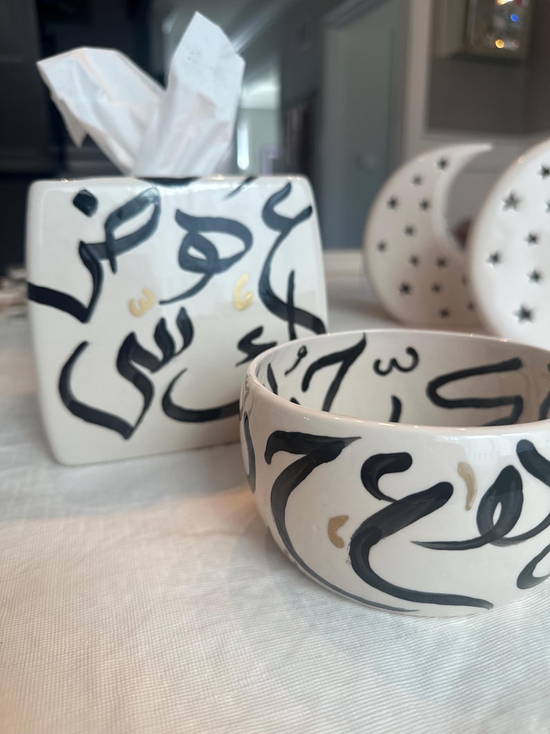 May include: A white ceramic tissue box and bowl set with black and gold calligraphy. The tissue box is rectangular with a square opening for tissues. The bowl is round. Two decorative moon-shaped objects are in the background.