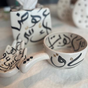 May include: A set of white ceramic home decor items with black and gold Arabic calligraphy. The set includes a tissue box cover, a small bowl with a handle, and a small dish. The items are on a white surface.