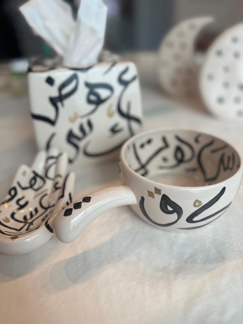 May include: A set of white ceramic home decor items with black and gold Arabic calligraphy. The set includes a tissue box cover, a small bowl with a handle, and a small dish. The items are on a white surface.