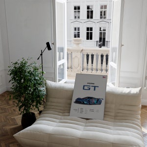 May include: A white poster with a blue and black Ford GT race car on it. The poster has the words "Ford GT" in black text at the top. The poster is sitting on a white couch in a room with a window.