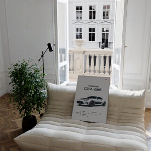 May include: A white poster with a black and white image of a Toyota GR-86 car. The poster is on a white couch in a room with a window overlooking a city street.