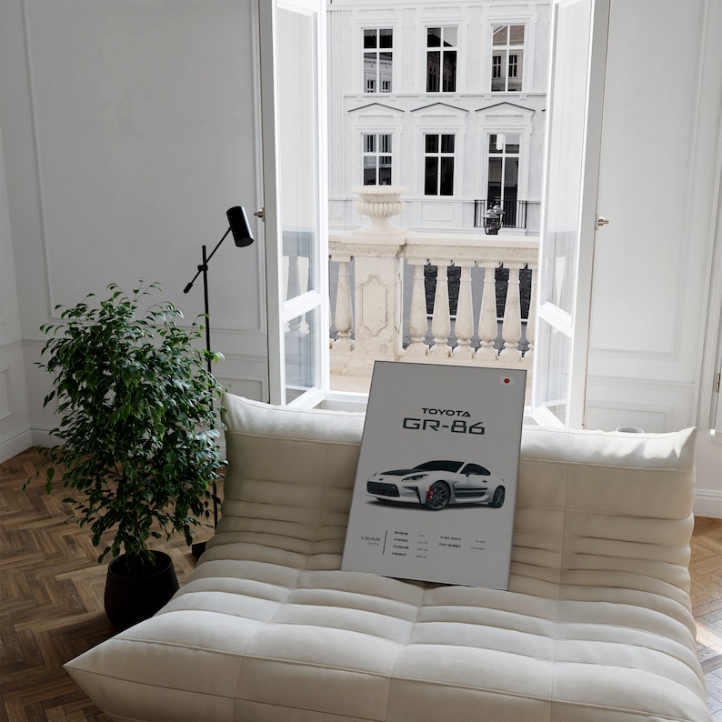 May include: A white poster with a black and white image of a Toyota GR-86 car. The poster is on a white couch in a room with a window overlooking a city street.