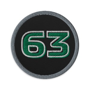 May include: A round, black patch with a grey border. The number 63 is embroidered in green with a white outline.