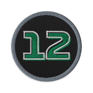 Può includere: Un cerotto rotondo nero con un bordo grigio. Il numero 12 è ricamato in verde con un contorno bianco.