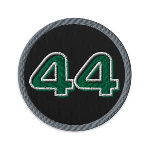 Könnte beinhalten: Runder, gestickter Aufnäher mit der Zahl "44" in Grün mit weißem Rand auf schwarzem Hintergrund. Der Aufnäher hat einen grau gestickten Rand. Das Design ist klar und auffällig, ideal für Kleidung oder Accessoires.