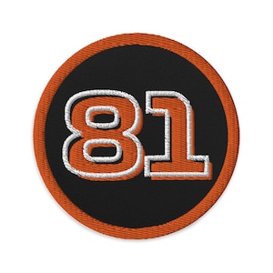 Puede incluir: Número 81 bordado en naranja y blanco sobre un círculo negro con borde naranja.