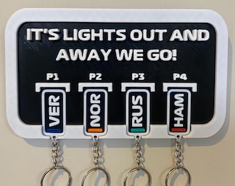 May include: A white wall-mounted key holder with the text "It's lights out and away we go!" and four key hooks labeled "P1 VER", "P2 NOR", "P3 RUS", and "P4 HAM".
