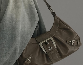 May include: A brown leather shoulder bag with silver buckles and a curved design. The bag has a short strap and a flap closure with a large buckle. The bag is worn over a denim jacket.