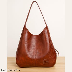 May include: A brown leather shoulder bag with a single strap. The bag has a curved shape and a small rectangular patch with text on the lower front. The bag is sitting on a wooden surface. The text "LeatherLofts" is at the bottom left.