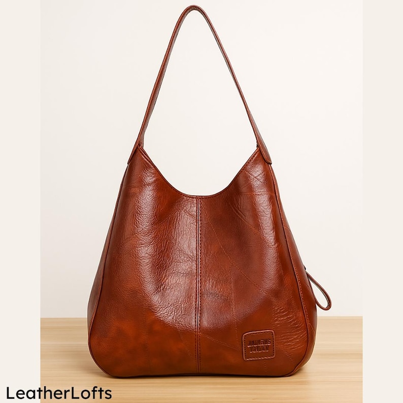 May include: A brown leather shoulder bag with a single strap. The bag has a curved shape and a small rectangular patch with text on the lower front. The bag is sitting on a wooden surface. The text "LeatherLofts" is at the bottom left.